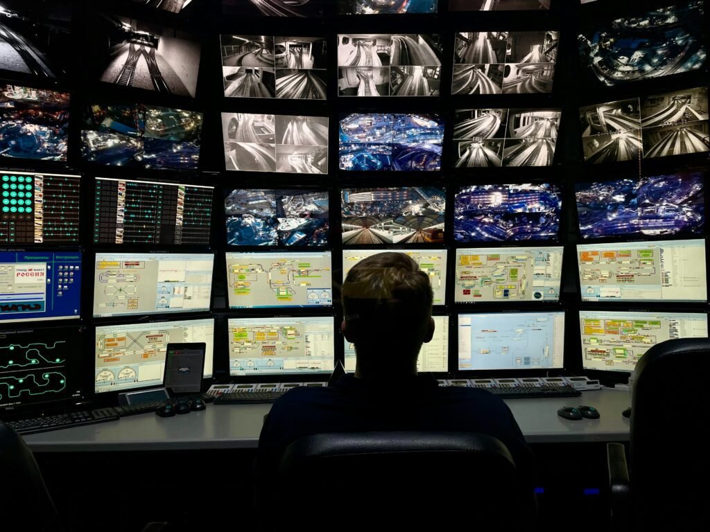 A person sits with their back to the viewer, facing a large wall of multiple monitors displaying various surveillance feeds and data interfaces, typical of a CCTV Monitoring Company.