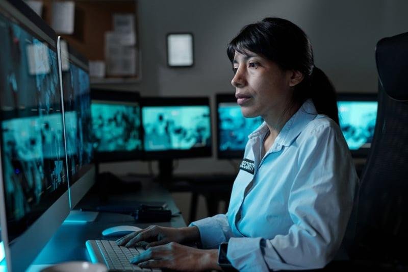 A focused female security officer monitoring multiple screens in a dark control room, representing a professional virtual doorman services provider ensuring reliable virtual doorman services for condo security Singapore