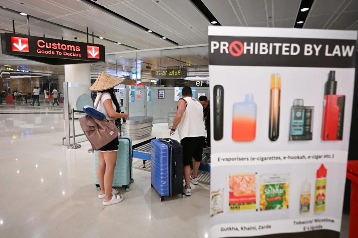A prominent "PROHIBITED BY LAW" sign at an airport customs checkpoint displaying an electronic cigarette and other smoking devices, warning travelers about strict Laws on Vapes enacted to protect youth safety.