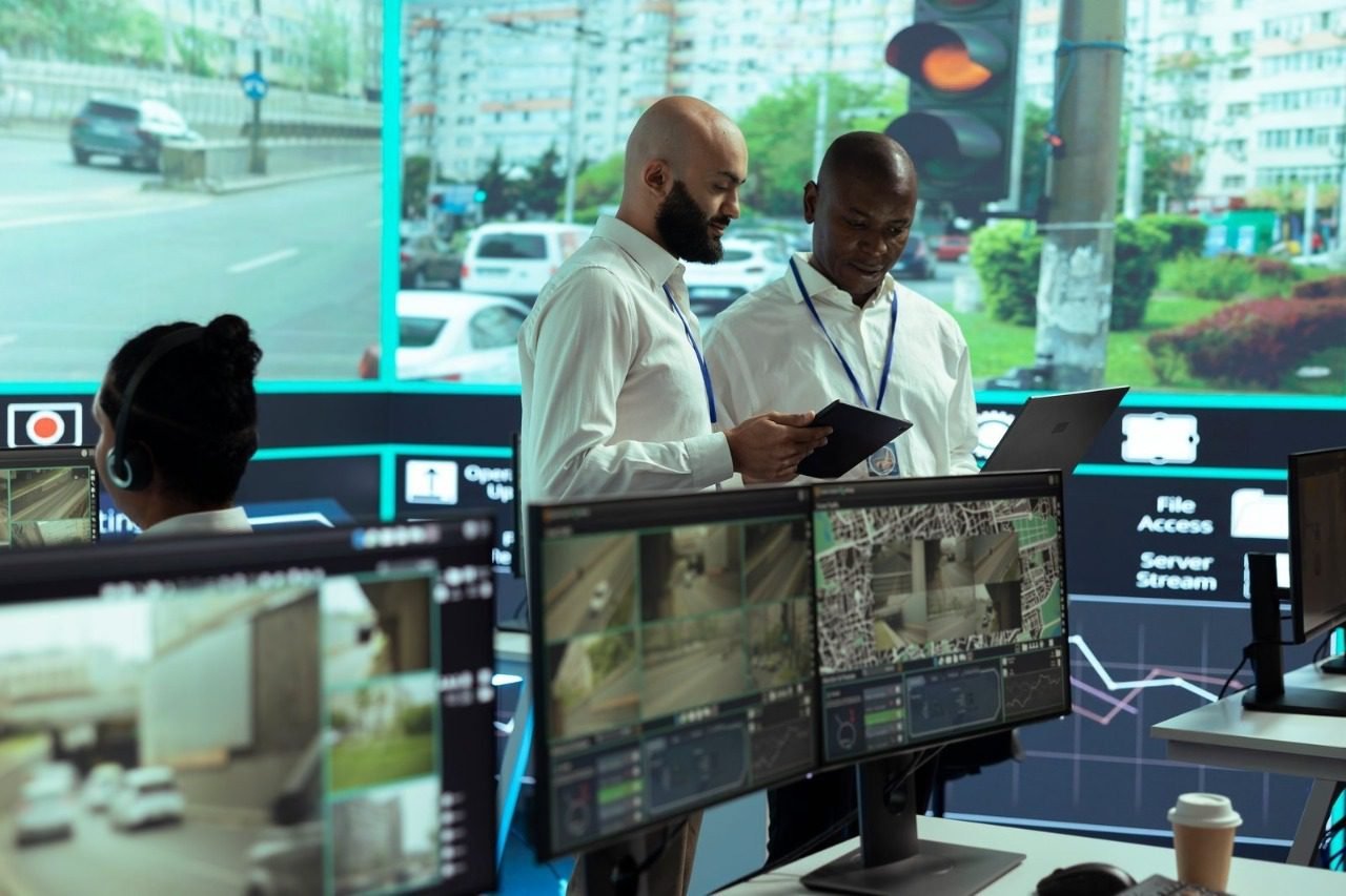 Two security professionals collaborate over a tablet in a state-of-the-art control room with massive video walls displaying live city traffic, illustrating the advanced infrastructure and expertise that factor into CCTV Monitoring Costs in Singapore for a reliable cctv monitoring service and security camera monitoring service.