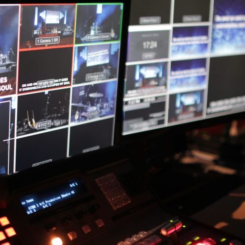 Close-up of a dark video production control console with brightly lit buttons and multiple screens displaying live camera feeds from a stage setup, resembling an operational 24/7 monitoring system.